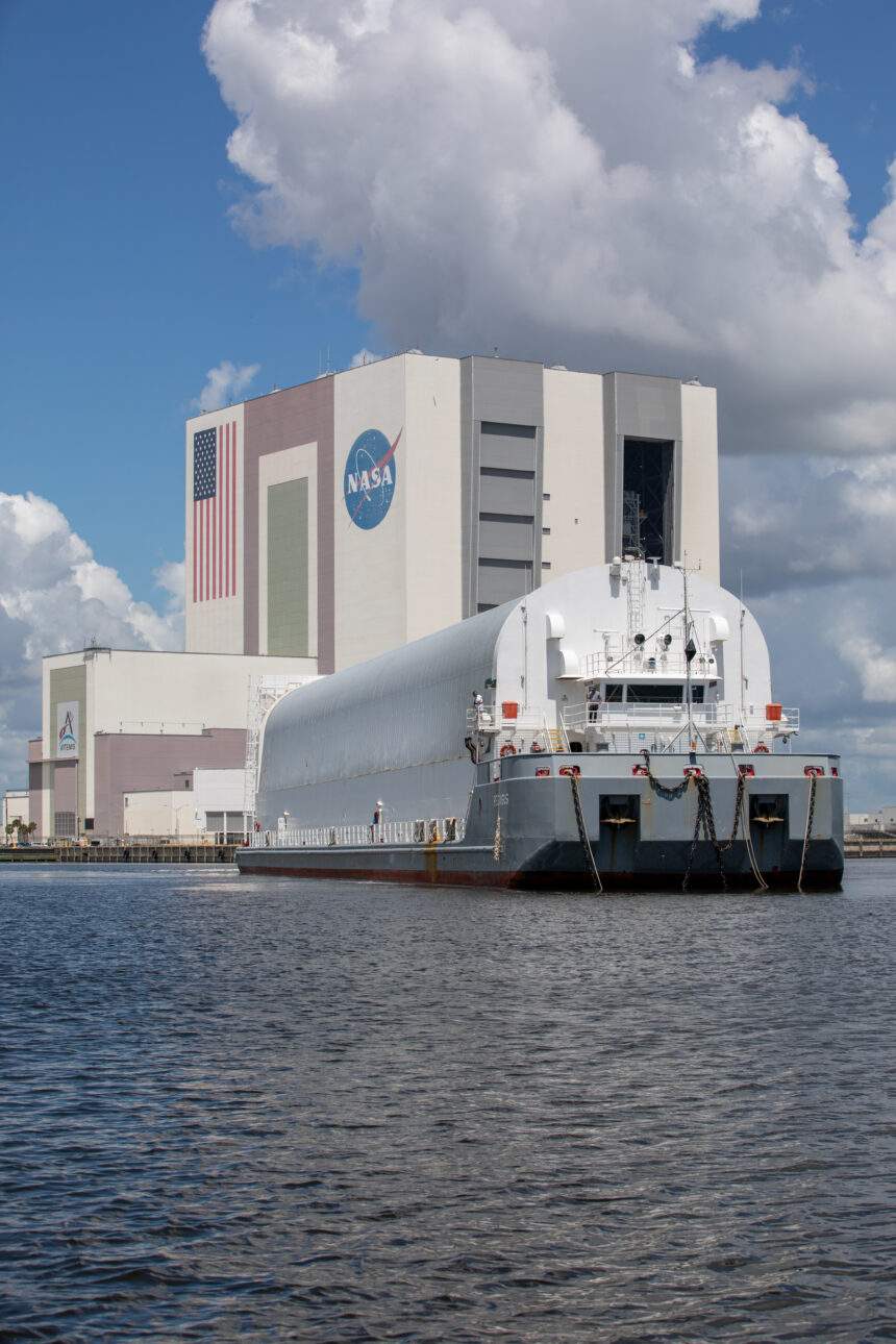 A large flat ship with a rounded structure on it approaches Kennedy Space Center's Vehicle Assembly Building. Dark blue water is in the foreground, while the ship moves toward the building in the background. The sky is blue, but huge, white fluffy clouds covering a good portion of it.