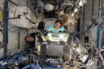 Two people look up at a camera. They each have their arms in gloves that go inside a lit-up rectangular box. We can see their arms through the windows on each side of the box. There are wires all around them inside the International Space Station.