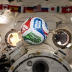 A white, red, blue, and green soccer ball floats inside the International Space Center. The FIFA logo is visible in the blue part of the ball facing the camera. The area in the background is mostly white, with a banner of country flags at the top of the photo.