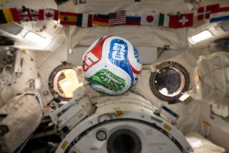 A white, red, blue, and green soccer ball floats inside the International Space Center. The FIFA logo is visible in the blue part of the ball facing the camera. The area in the background is mostly white, with a banner of country flags at the top of the photo.