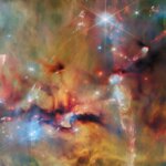 An area inside a star-forming molecular cloud. The background is covered with layers of gas and dust in blue, green and yellowish colors. Thicker clumps of cold dust, dark brown to black, block out light completely. Stars lie among and atop the clouds, from small orange ones to large white or blue ones. Waves and streams of glowing whitish gas are created by jets from protostars colliding with the surrounding material.