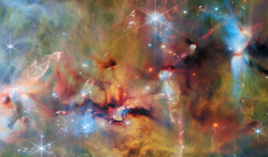 An area inside a star-forming molecular cloud. The background is covered with layers of gas and dust in blue, green and yellowish colors. Thicker clumps of cold dust, dark brown to black, block out light completely. Stars lie among and atop the clouds, from small orange ones to large white or blue ones. Waves and streams of glowing whitish gas are created by jets from protostars colliding with the surrounding material.