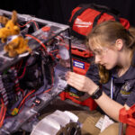 A student holds onto the edge of the rectangular body of a prototype lunar robot as she works on it. The rover has a rectangular body, which has multicolored attached to it. There is a small stuffed moose sitting on top of the rover.