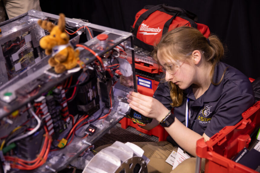A student holds onto the edge of the rectangular body of a prototype lunar robot as she works on it. The rover has a rectangular body, which has multicolored attached to it. There is a small stuffed moose sitting on top of the rover.