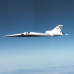 NASA’s X-59 quiet supersonic research aircraft flies through the blue sky. The plane has a distinctive long, sharp nose. There are mountains visible at the bottom of the photo.
