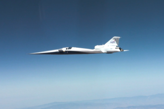 NASA’s X-59 quiet supersonic research aircraft flies through the blue sky. The plane has a distinctive long, sharp nose. There are mountains visible at the bottom of the photo.
