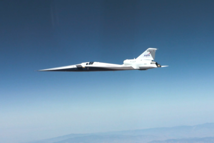 NASA’s X-59 quiet supersonic research aircraft flies through the blue sky. The plane has a distinctive long, sharp nose. There are mountains visible at the bottom of the photo.