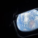 NASA astronaut Reid Wiseman looks out of the Orion spacecraft's rectangular window at Earth. He is barely visible in the dimly lit cabin, but Earth is bright. The entirety of Earth cannot be seen; in this view, Earth's round shape is cut off. Earth itself looks like a bright blue ball with swirling white clouds all over it; at center is a brown and green landmass.