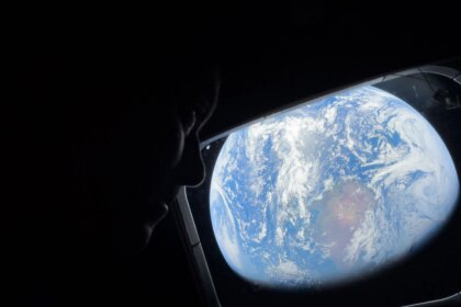 NASA astronaut Reid Wiseman looks out of the Orion spacecraft's rectangular window at Earth. He is barely visible in the dimly lit cabin, but Earth is bright. The entirety of Earth cannot be seen; in this view, Earth's round shape is cut off. Earth itself looks like a bright blue ball with swirling white clouds all over it; at center is a brown and green landmass.