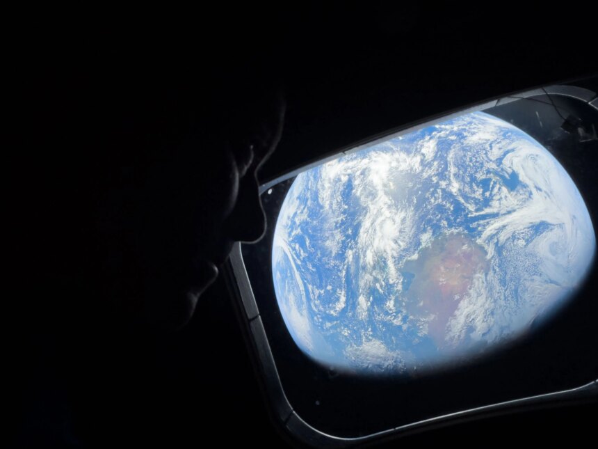 NASA astronaut Reid Wiseman looks out of the Orion spacecraft's rectangular window at Earth. He is barely visible in the dimly lit cabin, but Earth is bright. The entirety of Earth cannot be seen; in this view, Earth's round shape is cut off. Earth itself looks like a bright blue ball with swirling white clouds all over it; at center is a brown and green landmass.