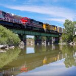 A train with many different colored cars crosses a green bridge over a body of water. There are trees along the shoreline.
