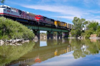 A train with many different colored cars crosses a green bridge over a body of water. There are trees along the shoreline.