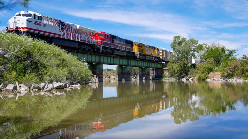 A train with many different colored cars crosses a green bridge over a body of water. There are trees along the shoreline.