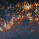 A large, long portion of one of the spiral arms in galaxy M51. Red-orange, clumpy filaments of gas and dust that stretch in a chain from left to right comprise the arm. Shining cyan bubbles light up parts of the gas clouds from within, and gaps expose bright star clusters in these bubbles as glowing white dots. The whole image is dotted with small stars. A faint blue glow around the arm colors the otherwise dark background.