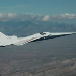 NASA’s X-59 flies above the Mojave Desert on a clear day. The white aircraft has light gray, red, and blue accents, with a NASA logo and the number 859 on its tail. It appears flying level over the desert landscape, with a mountain range visible on the horizon and a trail of clouds above.