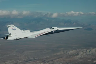 NASA’s X-59 flies above the Mojave Desert on a clear day. The white aircraft has light gray, red, and blue accents, with a NASA logo and the number 859 on its tail. It appears flying level over the desert landscape, with a mountain range visible on the horizon and a trail of clouds above.